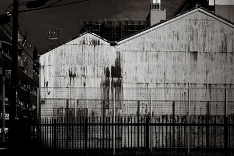 A stained corrugated warehouse wall with twin peaked roofs rises behind metal fencing in black and white.