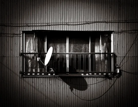 A satellite dish projects from a window balcony on a corrugated wall in a black and white view.