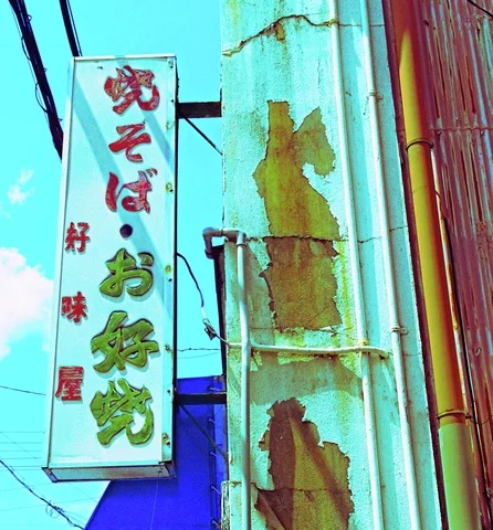 A narrow Japanese sign projects beside peeling turquoise siding beneath overhead wires and bright blue sky.
