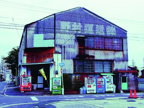 Vending machines line a heavily weathered industrial corner building with broad faded upper lettering.