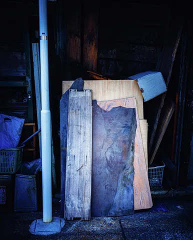 Several rough wooden boards lean beside a pale drainpipe in blue-toned shadows.