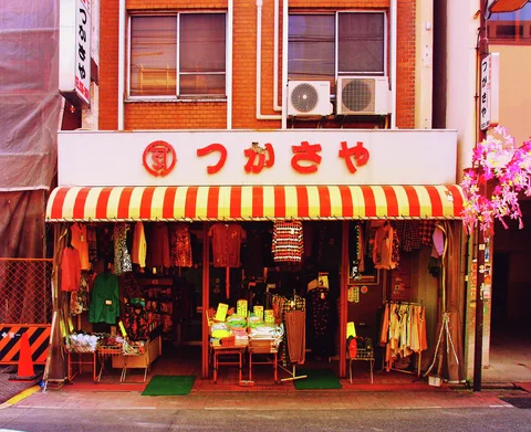 Clothing and merchandise fill an open shop beneath a red and white striped awning and orange Japanese lettering.