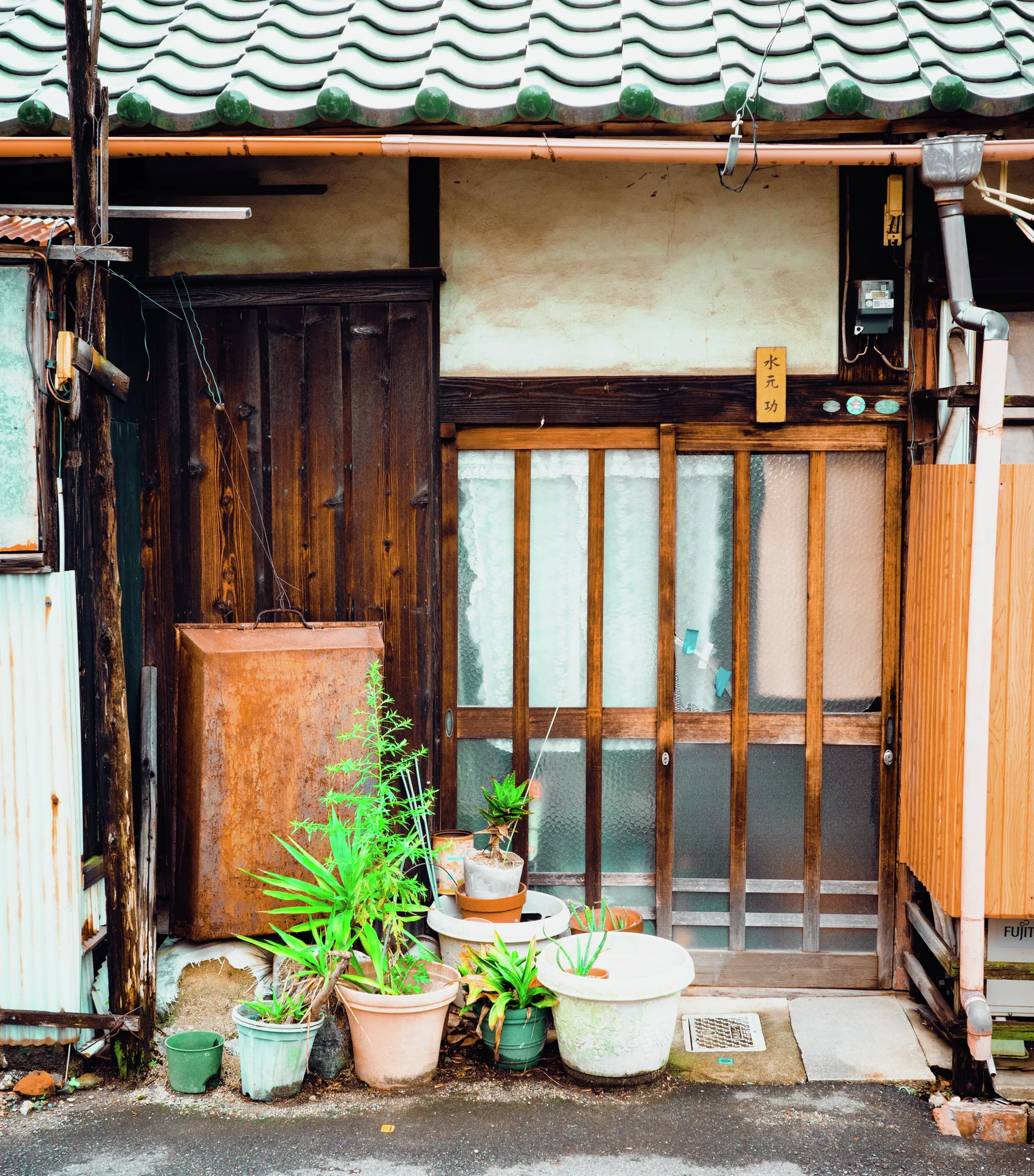 Potted plants line the entrance of a weathered wooden house beneath a green tiled roof.