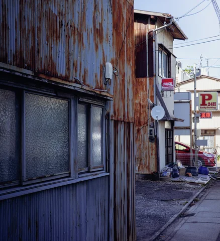 Rust streaks cover corrugated metal walls above frosted windows beside a narrow lane and overhead wires.