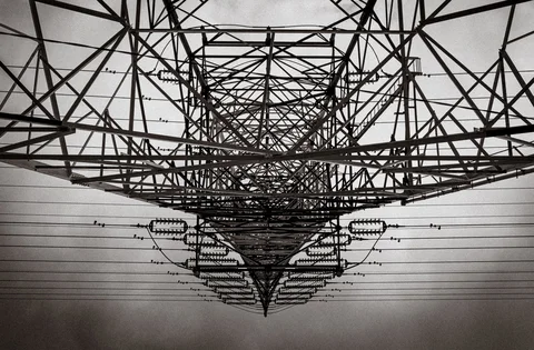 A dense lattice of electricity tower beams converges overhead among parallel power lines in black and white.
