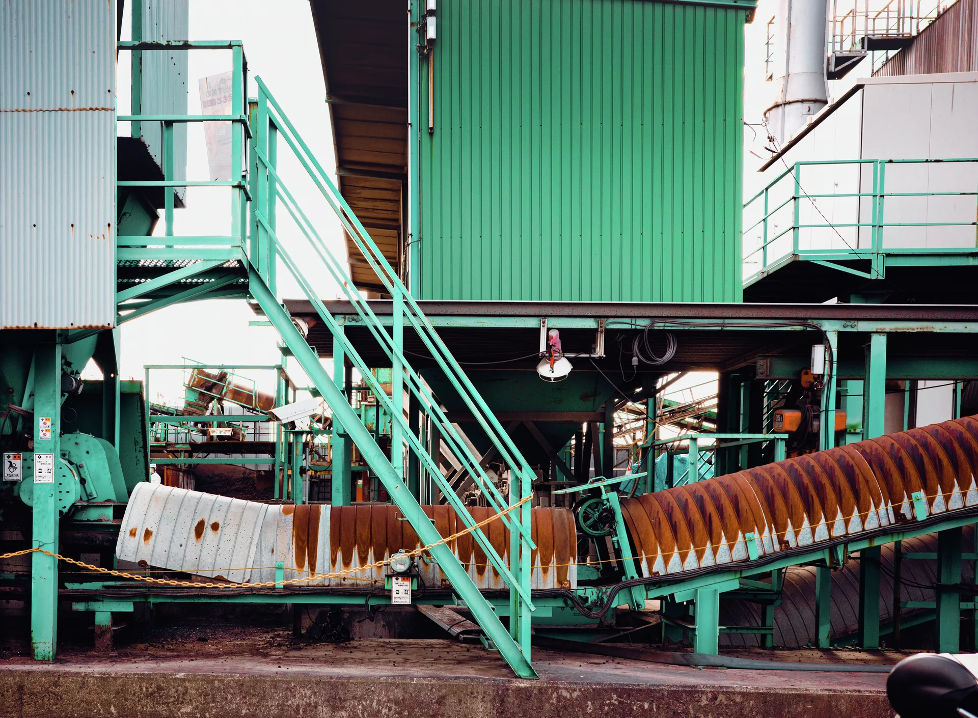 Turquoise stairs rise beside corrugated industrial walls above a rust-striped curved conveyor structure.