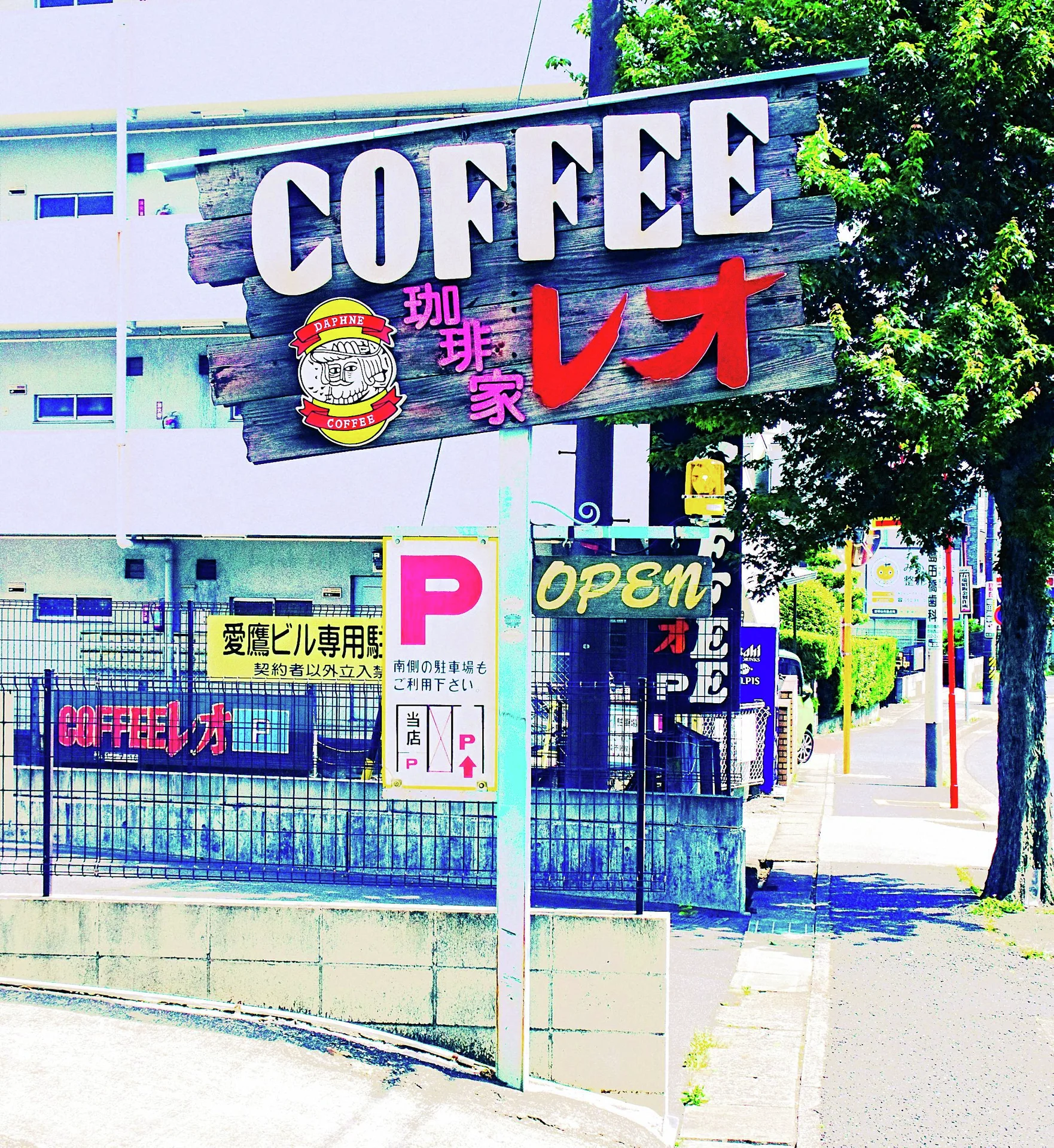 A large coffee sign stands above a street corner with a parking sign and leafy tree.