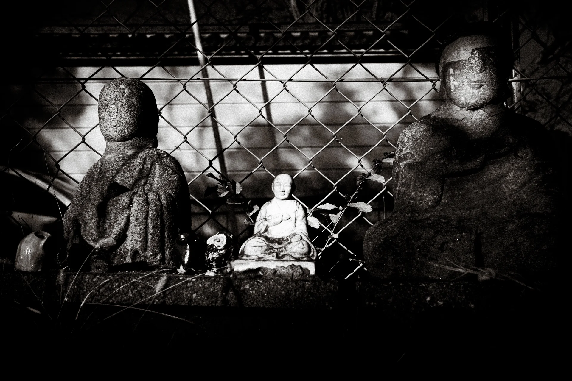 Three seated stone figures of differing sizes sit before a chain-link fence in black and white.