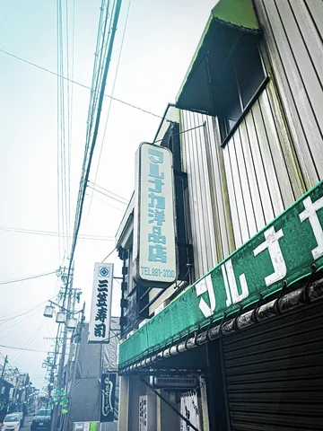 A vertical Japanese sign hangs beside a pale storefront above a green fascia and closed shutter.