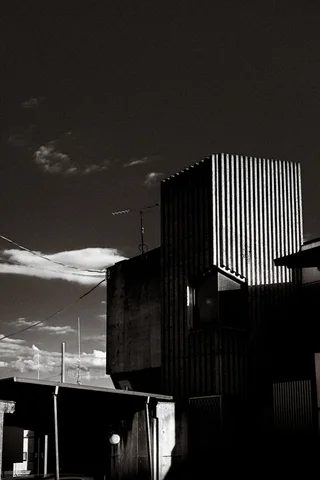 A tall corrugated industrial building rises beneath a dark sky and bright clouds in monochrome.