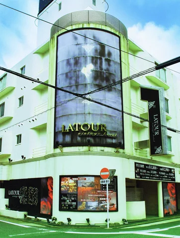 A rounded glass tower rises above a corner storefront beneath blue sky and crossing wires.