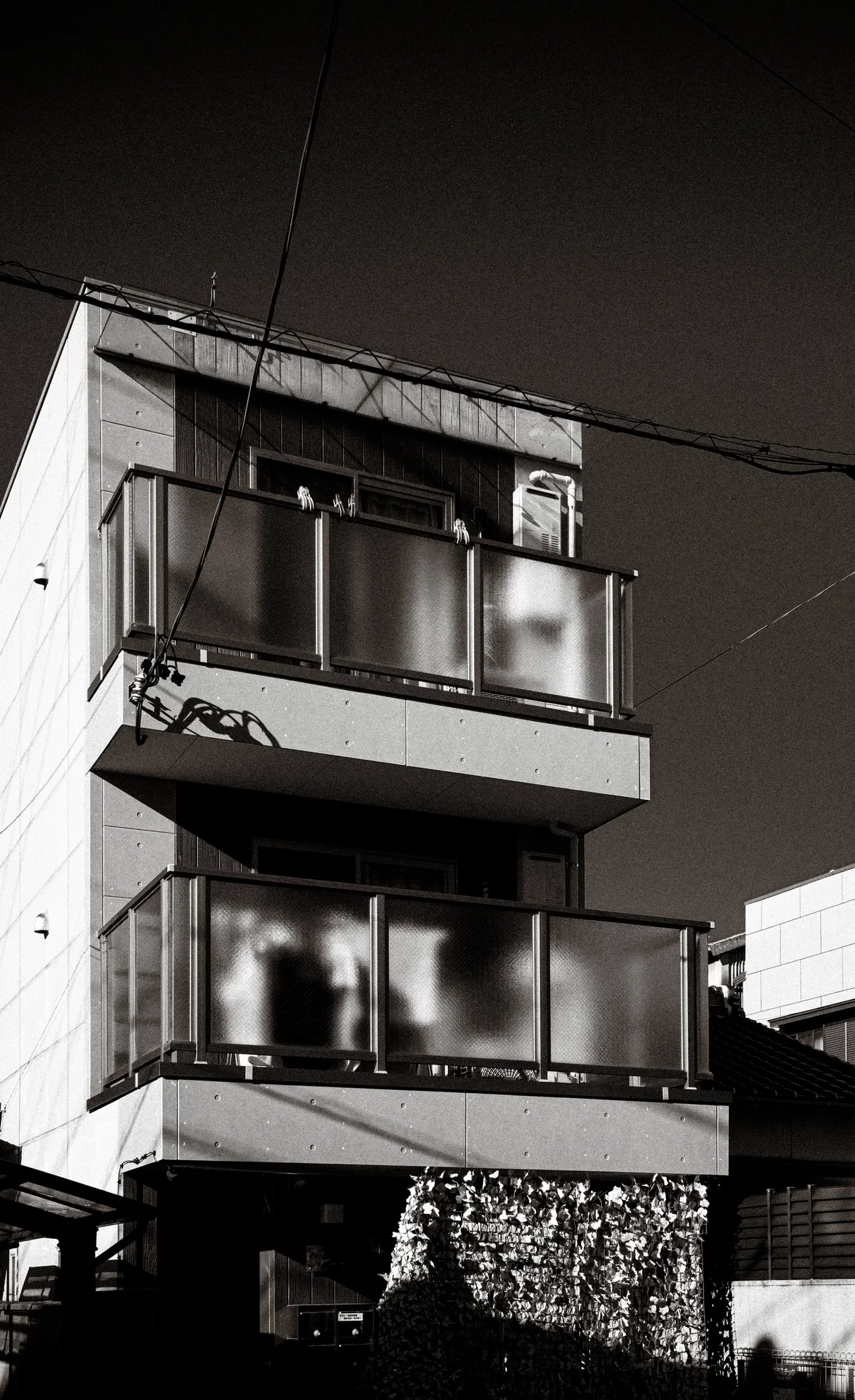 Two projecting balconies with translucent panels catch sunlight on a narrow building in black and white.