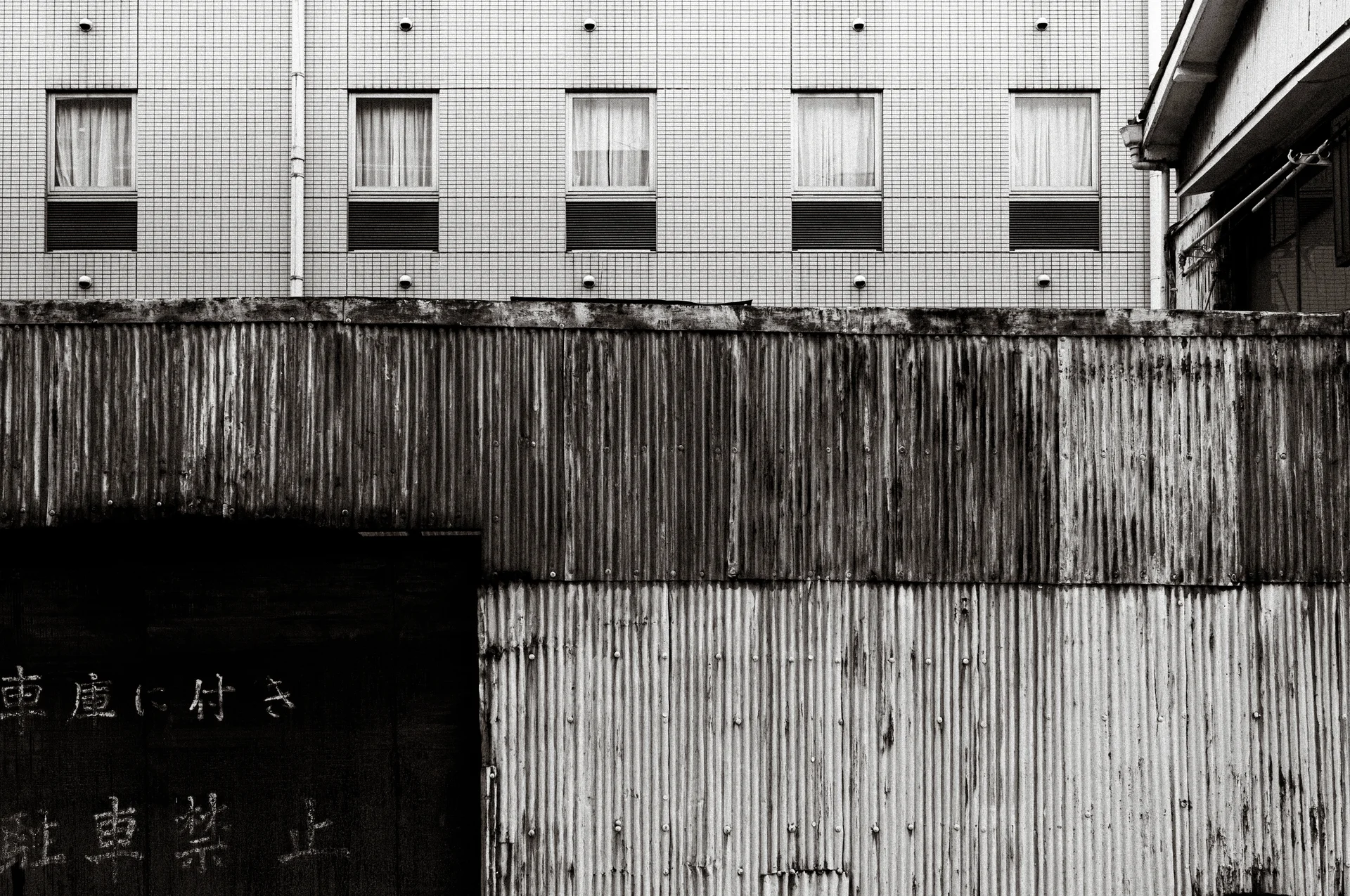 Weathered corrugated metal walls stand below rows of windows in a black and white urban view.