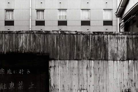 Weathered corrugated metal walls stand below rows of windows in a black and white urban view.