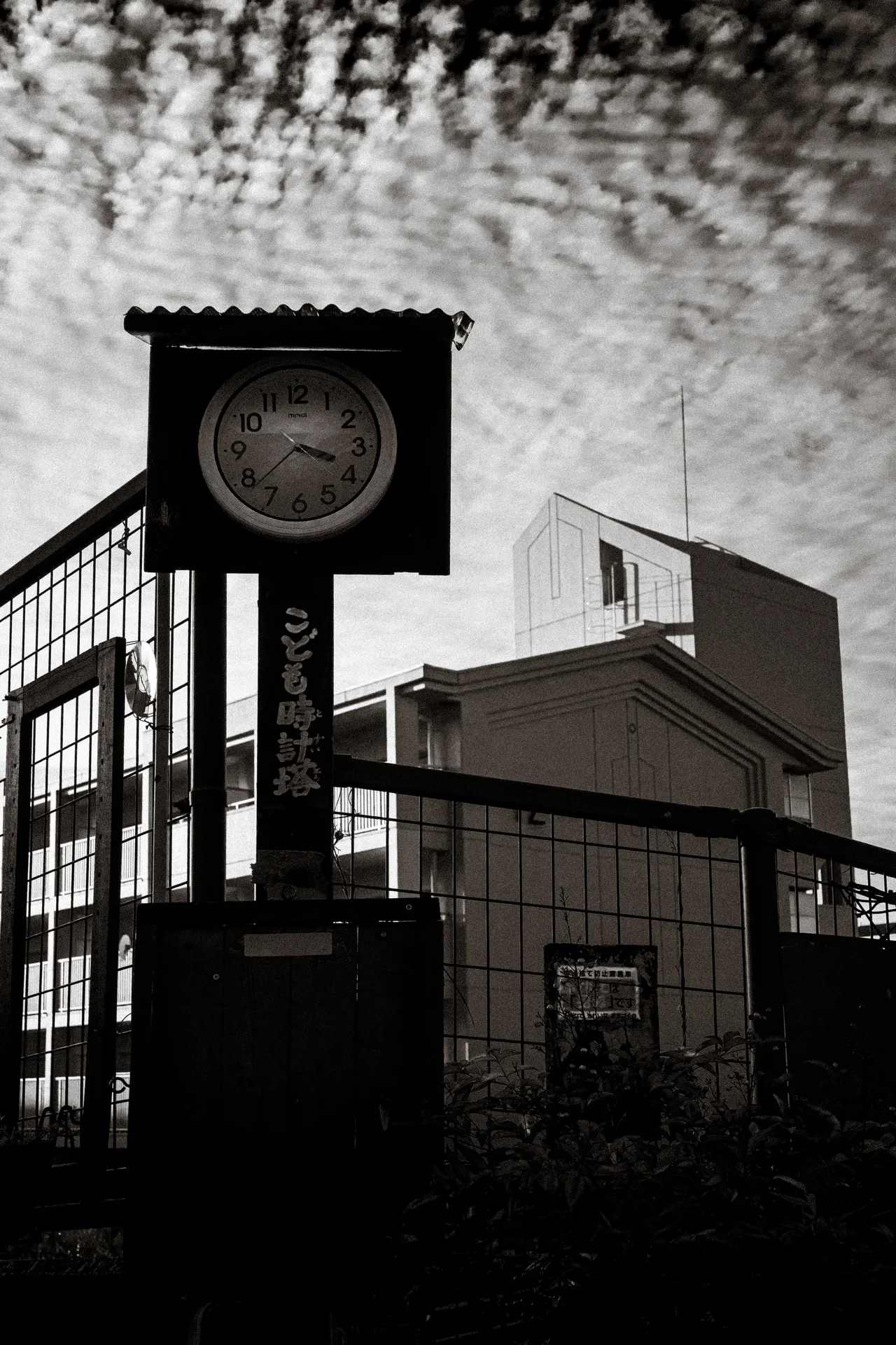 A round clock hangs within a square frame above a wire fence and buildings in black and white.