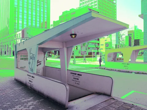 A pale covered bench structure stands beside a broad roadway and tall city buildings.