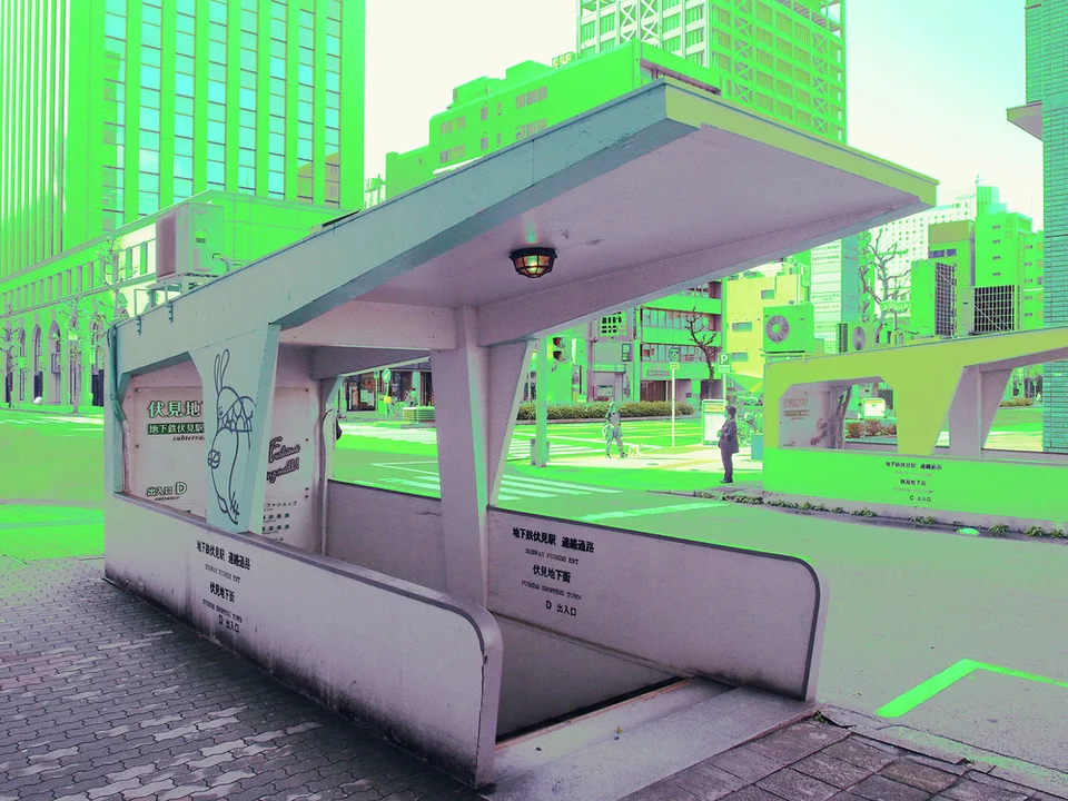 A pale covered bench structure stands beside a broad roadway and tall city buildings.