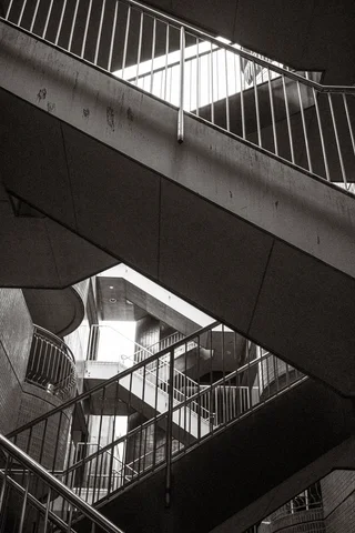 Concrete stair flights and metal railings intersect beneath a bright opening in black and white.