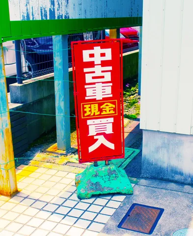 A red vertical sign with bold pale characters stands on a green base beside a tiled sidewalk.