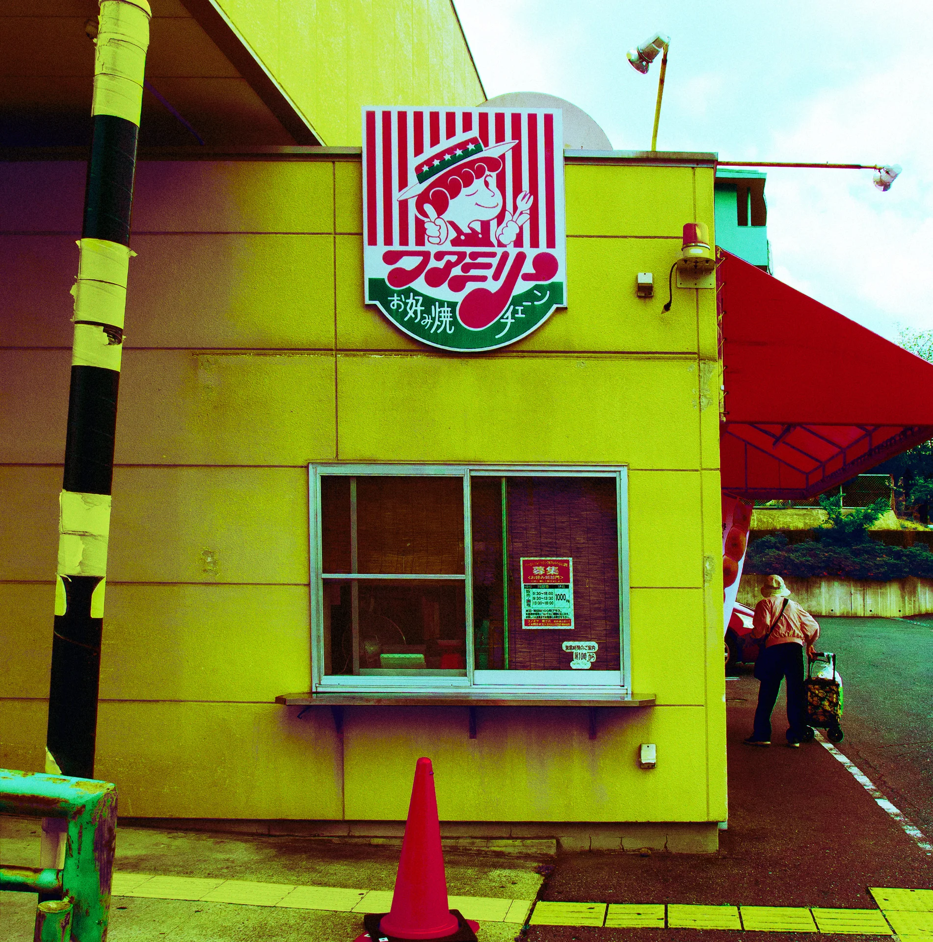 A striped illustrated sign hangs above a window on a yellow building beside a red canopy and orange cone.