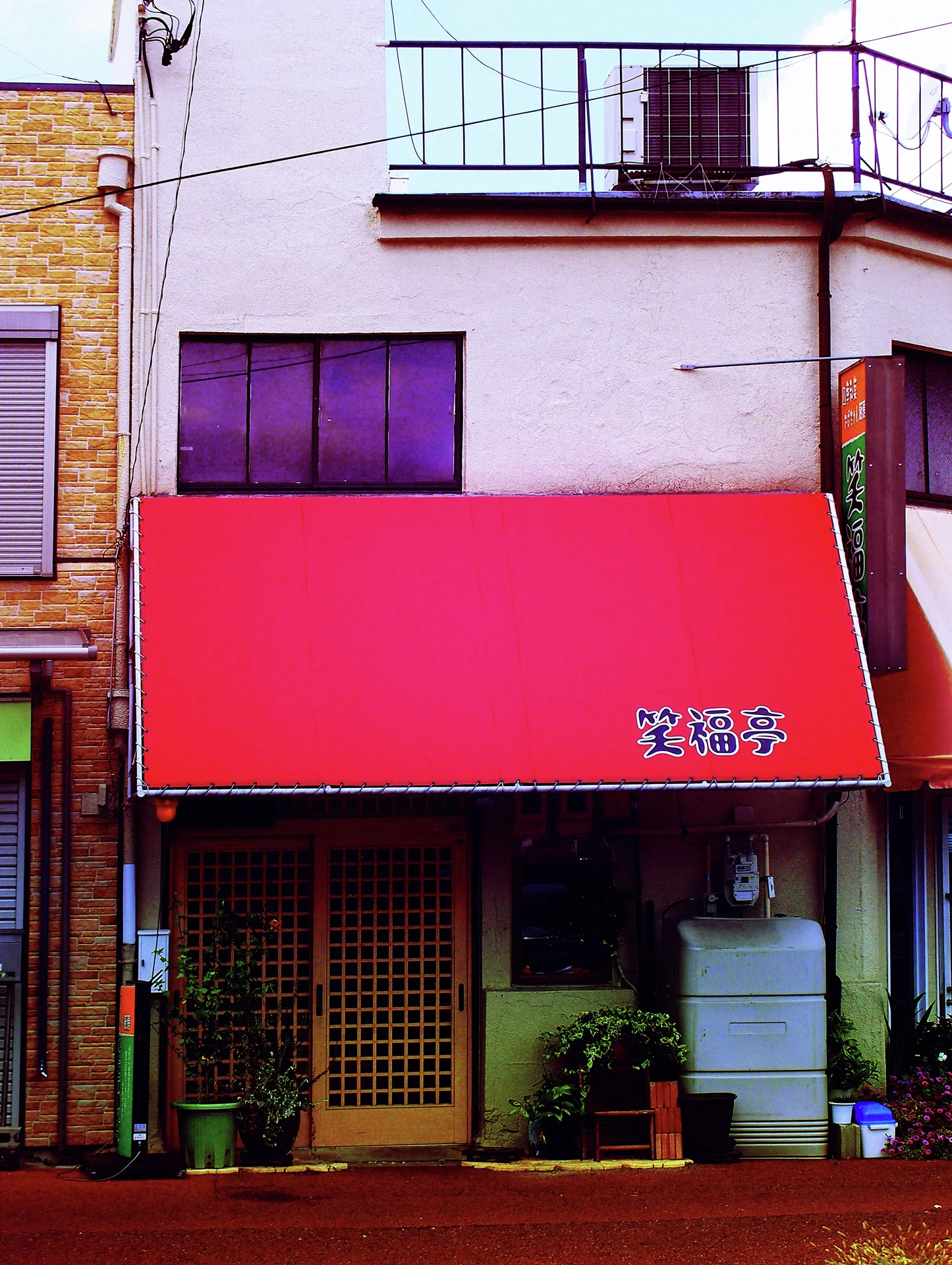 A vivid pink awning shades a narrow storefront beneath a pale upper wall and rooftop railing.