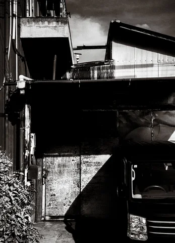 Sharp shadows divide a cramped gap between weathered buildings and a parked car in black and white.