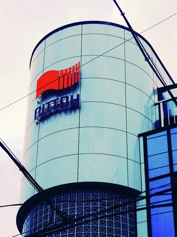A cylindrical pale building corner displays a red emblem above dark lettering behind diagonal utility wires.