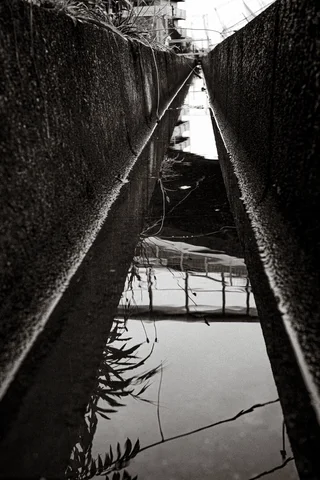 Still water reflects overhead structures within a narrow concrete drainage channel in a black and white view.