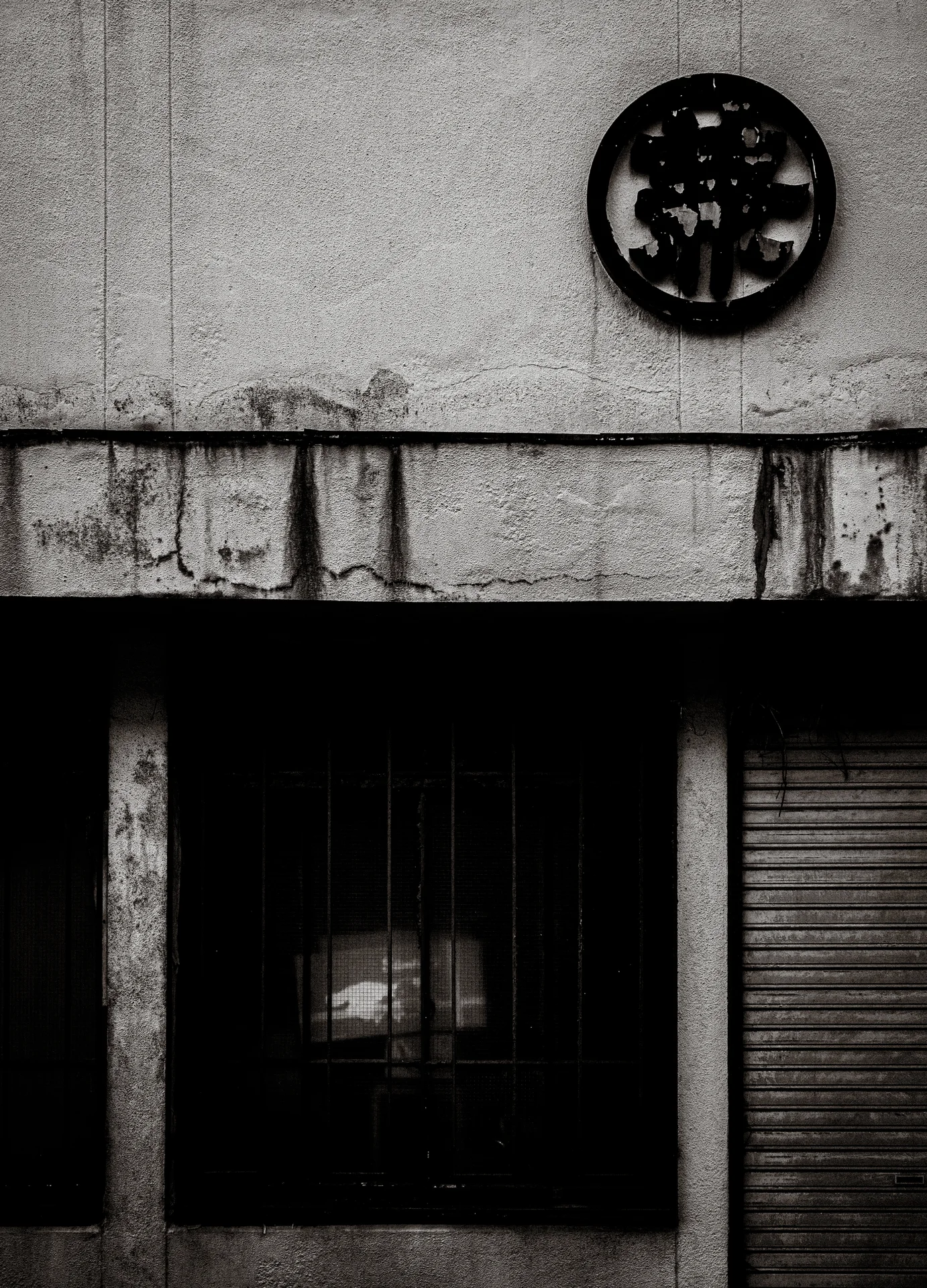 A circular wall emblem hangs above a dark recessed doorway and closed shutter in black and white.