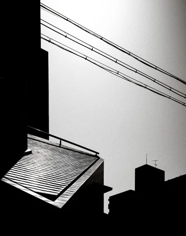 Diagonal overhead wires cross a pale sky above silhouetted rooftops and a sunlit corrugated awning.