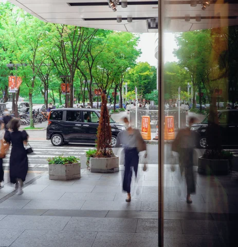 Blurred pedestrians pass glass doors beside potted trees, with a dark van visible across the sidewalk.