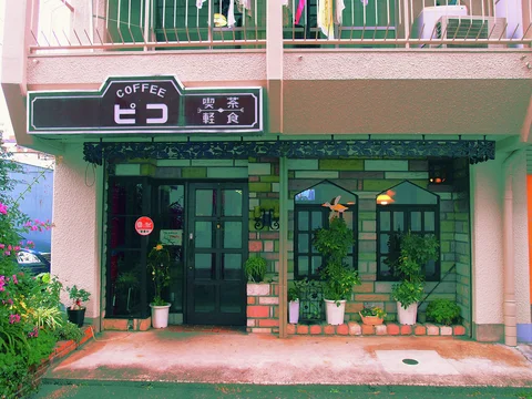A cafe entrance has dark grid-patterned glass doors, brick trim, and tall shrubs in pale pots.