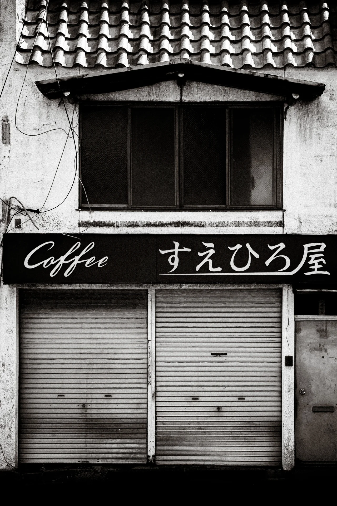 A Coffee sign with Japanese lettering spans closed shutters beneath a tiled roof in black and white.