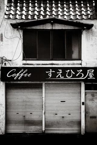 A Coffee sign with Japanese lettering spans closed shutters beneath a tiled roof in black and white.
