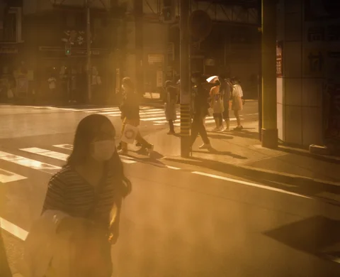 A woman stands near a sunlit crossing while pedestrians and their long shadows extend behind her.