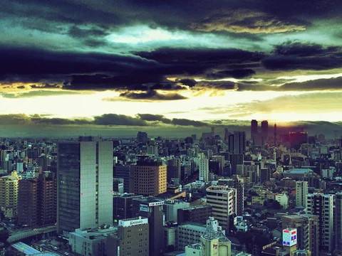 Dense city buildings stretch beneath dark layered clouds and a bright yellow strip along the horizon.