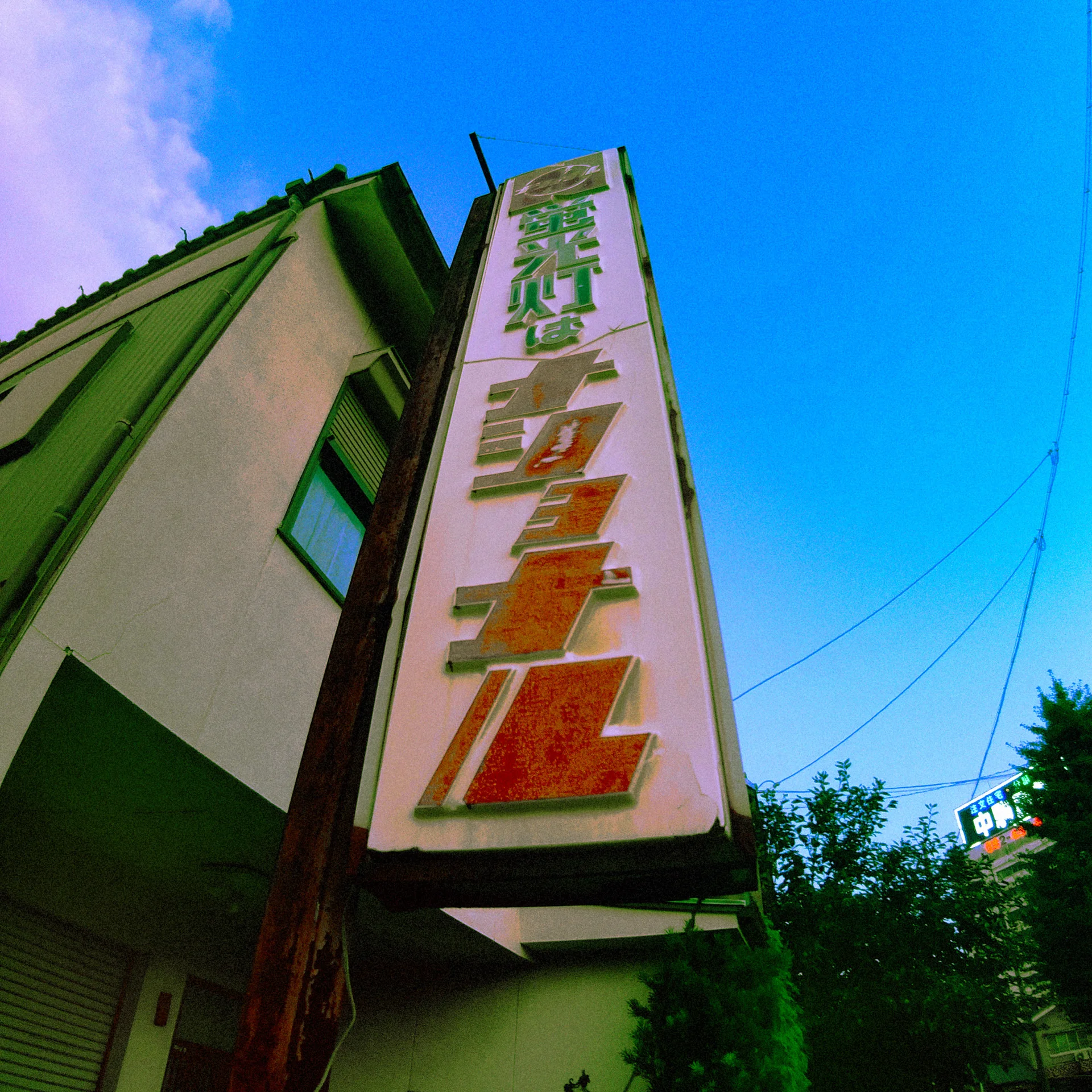 A tall faded vertical sign projects from a pale building into bright blue sky.