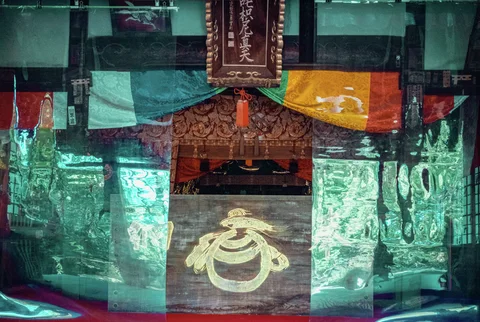 Turquoise translucent panels frame a carved wooden altar beneath multicolored fabric and a hanging Japanese plaque.