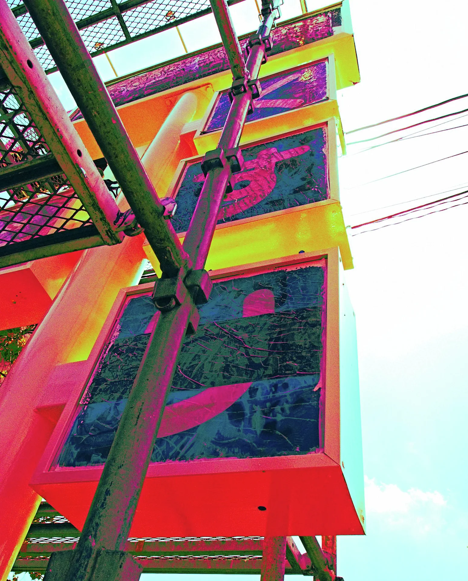 A red-and-yellow building rises behind diagonal metal framework with dark rectangular windows and overhead wires.