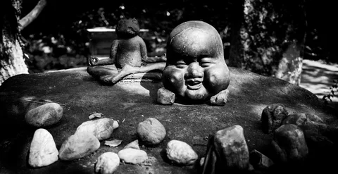 A smiling sculpted head rests among stones, with a cross-legged frog figure behind it in black and white.