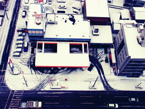 An overhead view reveals snow-covered flat rooftops, parked cars, and dark curving tracks across a central lot.