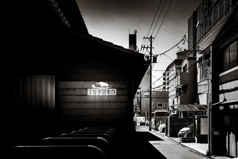 A narrow street passes a dark wooden building and taller facades beneath overhead wires.