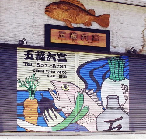 A colorful fish and vegetables decorate a closed shutter beneath a large wooden fish mounted on the wall.