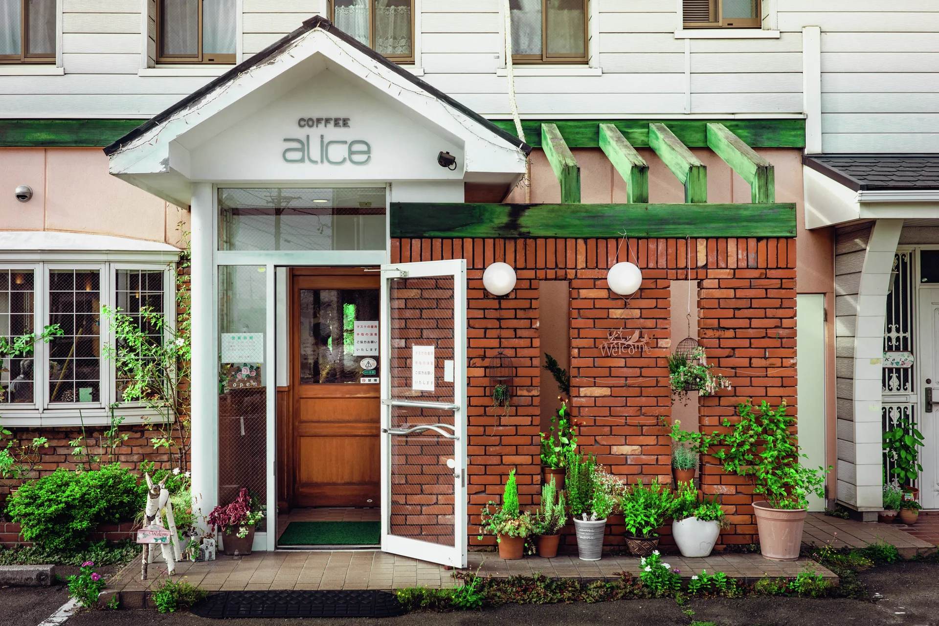 A small coffee shop has a peaked white entrance, brick walls, and abundant potted plants.