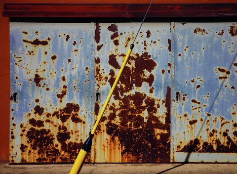 Heavy brown rust mottles a pale blue metal door behind a diagonal yellow support cable.