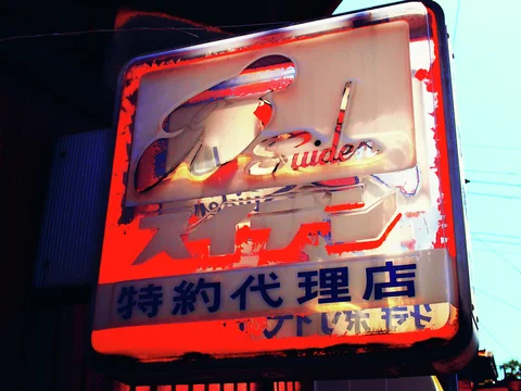 A peeling red and cream projecting sign bears stylized lettering and dark blue Japanese text.