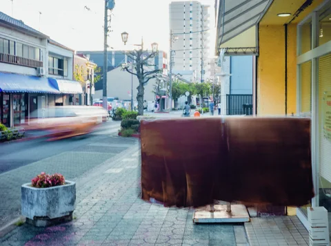 A large rusty panel stands beside a yellow storefront while a blurred vehicle passes a flower planter.