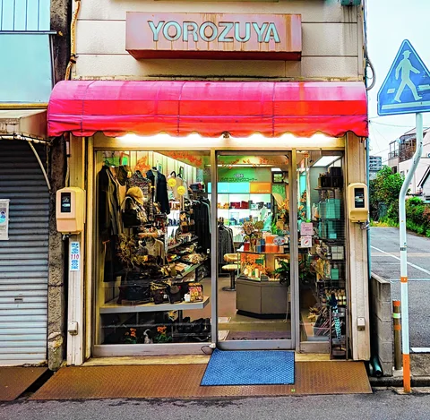 A red awning shades an open shop filled with displayed objects beside a blue pedestrian crossing sign.