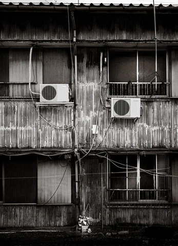 Two air conditioners hang between windows on a weathered wooden facade in black and white.