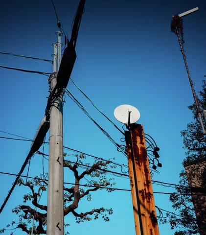 A white satellite dish tops a weathered wooden pole beside utility wires against blue sky.